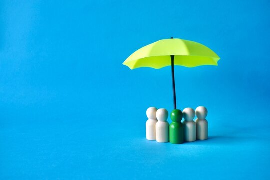Wooden peg figures with a green leader figure standing under a small yellow umbrella against a vibrant blue background, representing leadership, team protection, and group insurance concepts.