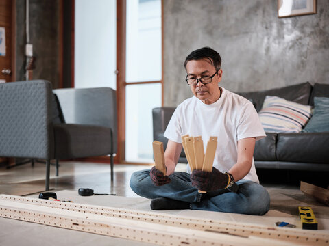 Asian mature man sitting and preparing flat-pack furniture parts and DIY equipment for self-assembly, home improvement, wooden craft installation, hobby workshop, carpentry handyman active lifestyle.