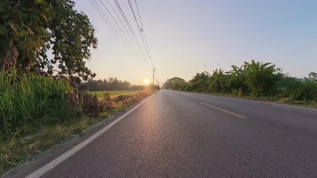 Scenic rural road during golden hour, with warm sunlight reflecting on the asphalt, lush green vegetation, utility poles and distant mountains beneath a clear sky.