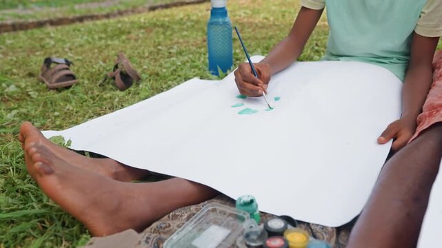 Children sit outdoors painting together. One applies green paint to white canvas while feet rest on grass supplies like paints and water bottle are visible