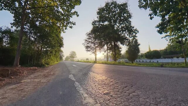 Scenic rural road lined with trees during golden hour, with warm sunlight shining through the foliage and reflecting across the asphalt surface.