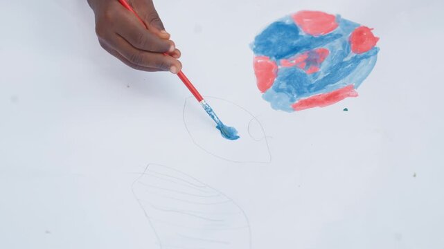 Close up view of hands using red handled brushes with light blue and dark blue watercolor paints to fill in pre drawn outlines on white paper. scene focuses solely on painting process
