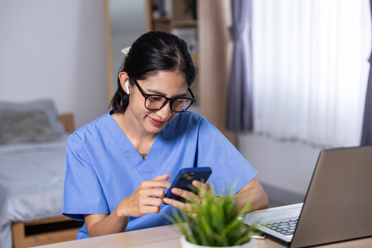 Asian nurse in blue uniform using smartphone beside laptop at home