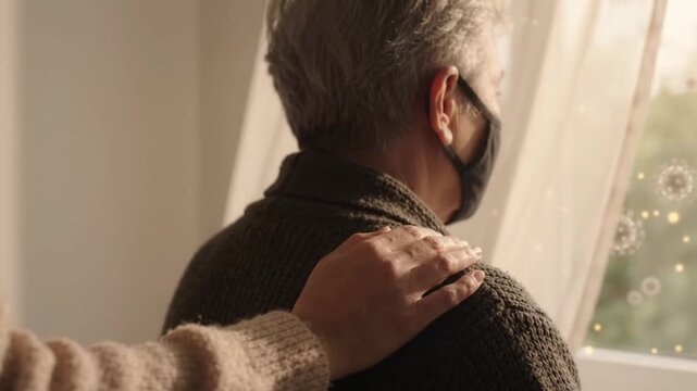 Elderly person wearing face mask looks out window with virus particles visible while supportive hand gently touches shoulder showing care health and virus prevention during challenges