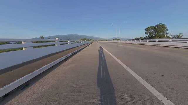 Cyclist shadow stretching across a bridge during a scenic ride, with distant mountains, road markings and clear blue sky creating a peaceful outdoor cycling scene.