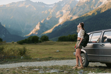 Naklejka premium Woman traveler beside a car in a mountainous landscape, backpack ready, gazing at distant peaks, outdoor adventure, nature scenery, bright daylight, calm mood.