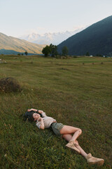Naklejka premium Calm portrait of a woman resting in a meadow beneath distant hills, enjoying outdoor space, sunlit grass and tranquil scenery, a quiet moment of leisure and nature.