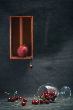 Surreal still life with red apple in wooden box and falling cherry into wine glass with spilled cherries on dark background