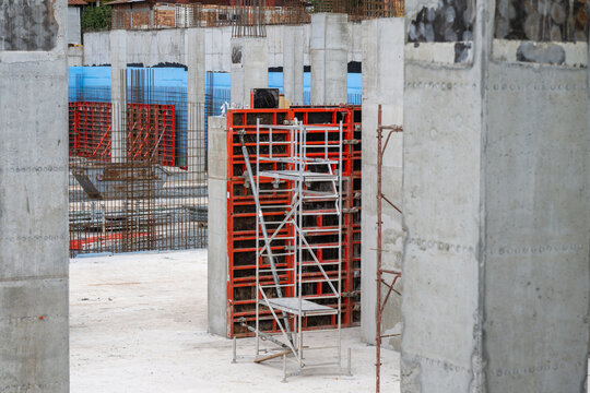 A ground-level view of a building construction site showing reinforced concrete columns, modular red wall formwork, and an aluminum scaffolding tower standing on a concrete foundation slab.