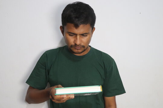 Young man holding and reading a closed book wearing a green t shirt against a white wall