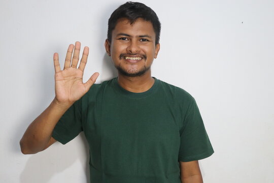 Young man in a green t shirt waving hand with a cheerful smile against white wall
