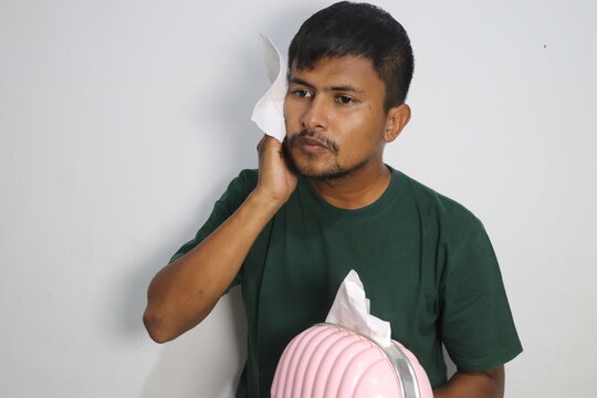 Young man holding tissue paper against his cheek while standing against a white wall