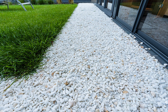 View along narrow bed of white decorative gravel bordering dark framed glass patio doors, with dense green lawn beside it, cool daylight and sharp texture contrast for exterior garden design.