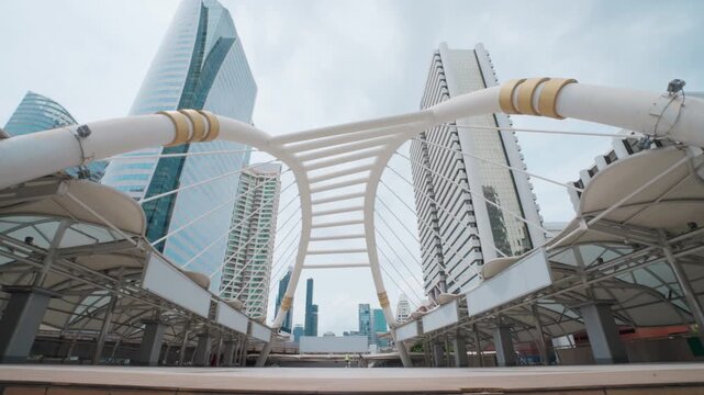Modern arch bridge spans urban skyline. Steel beams curve elegantly upward toward sky. Glass towers reflect daylight in distance