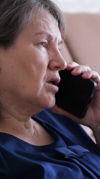 Woman talks on smartphone during quiet phone conversation. Older woman remains seated indoors, holding handset close and alternating between listening and speaking with serious look.