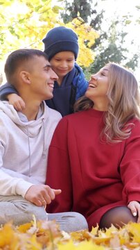 Happy parents sit with young child outdoors, smiling and sharing affectionate kisses among autumn leaves. Family enjoys tender moment together in warm fall light beneath golden trees.