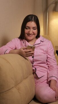 Smiling young woman in cozy pajamas relaxing on a sofa in a warm living room, happily browsing social media, texting friends, and using mobile applications on her modern smartphone