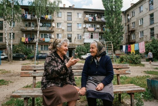 Two older women chatting on a weathered bench in a courtyard of a Soviet-era apartment block