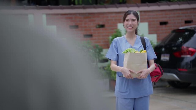 A healthcare worker arrives home holding a paper bag of fresh groceries, smiling warmly after returning from work and greeting family at the house entrance.