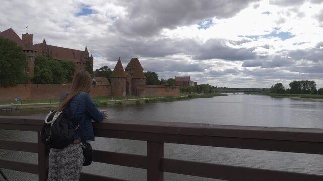 Female traveler with a backpack leans on a bridge railing and gazes at the red brick Teutonic Malbork Castle on the Nogat River under a cloudy summer sky in Poland.