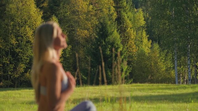 Caucasian blonde woman in bokeh sits outdoors with hands on her chest, breathing deeply in a calming mindfulness practice connecting with nature, with a sunlit forest visible behind her.