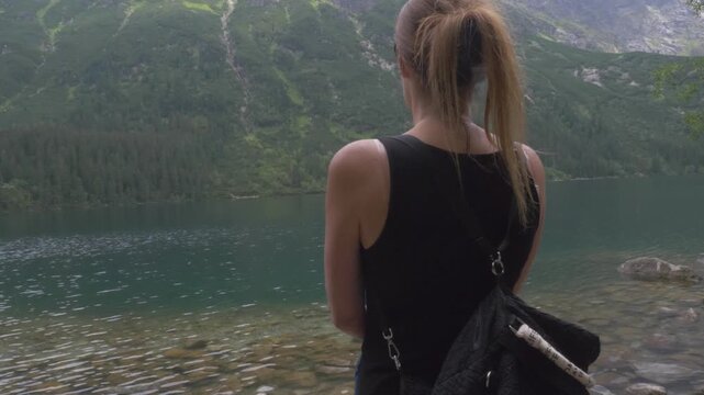 Hiker woman with a backpack walks to the transparent water of Morskie Oko lake and looks at the forested slopes of the Tatra Mountains in Tatra National Park.