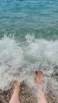 Sea wave splashing over bare feet on a pebble beach. The video represents concepts of summer vacation, relaxation and seaside lifestyle.