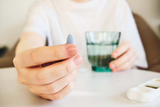 Person holding single blue pill, preparing to take medication with glass of water