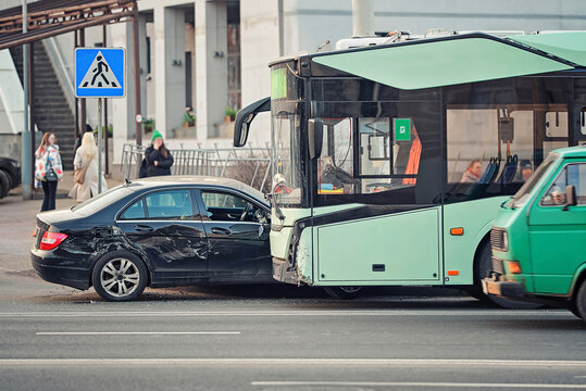 Collision between black car and green public transport trolleybus on city road, crumpled car side and bus front, with bystanders watching from sidewalk, traffic accident aftermath. Bus crash with car