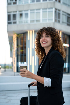 Young business white woman holding coffee and pulling suitcase