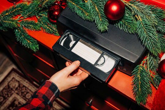 female hand inserts a Christmas-themed VHS tape into a VCR decorated with fir branches and a red bauble, ready to watch nostalgic home videos from the 90s