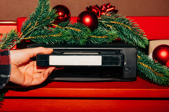female hand inserts a Christmas-themed VHS tape into a VCR decorated with fir branches and a red bauble, ready to watch nostalgic home videos from the 90s