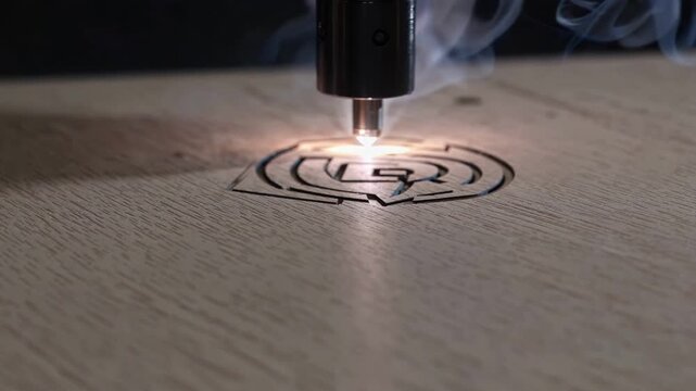 Computer controlled laser carver etches detailed symbol on wooden plate with pinpoint beam. Industrial engraving tool works across smooth timber surface, generating vapor and steady glow.
