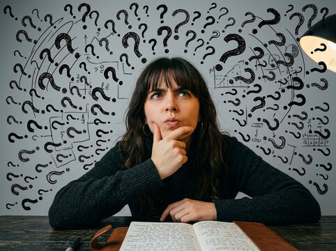 Thoughtful young woman sitting at desk with many question marks on