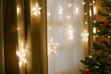 Fototapeta premium Luminous star shaped fairy lights hanging on window beside decorated Christmas tree indoors. Holiday background suitable for winter season marketing and Christmas party event promotions