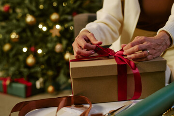 Fototapeta premium Cropped hands of Caucasian adult wrapping holiday present with ribbon near Christmas tree. Suitable for seasonal retail marketing, festive gift giving campaigns, greeting concepts