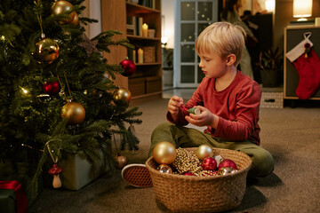 Fototapeta premium Caucasian child decorating Christmas tree with ornaments at home, celebrating winter holidays with family. Useful for seasonal marketing campaigns and holiday promotions