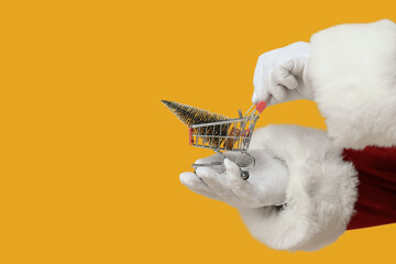 Fototapeta premium Christmas shopping and holiday retail sales, Santa Claus holding a miniature shopping cart with a small pine tree on a solid yellow background