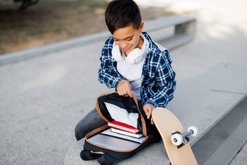 Schoolboy organizing books in backpack with skateboard outdoors © djile