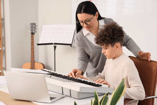 Music education and private tutoring, teacher helping young boy learn to play electronic keyboard in classroom with laptop and sheet music
