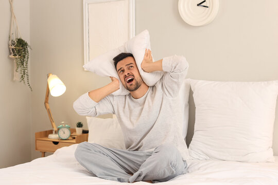 Insomnia and sleep deprivation, stressed man covering ears with pillow in bedroom to block noise and find peace during mental health struggle