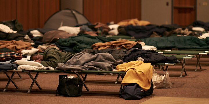 A room filled with cots set up as a temporary shelter, with sleeping bags and personal belongings scattered around, showing a scene of homelessness or displacement.