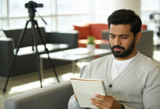 Focused Indian man writing in notebook sitting in modern studio with camera tripod in background for content creation