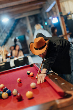 Person wearing a cowboy hat leans over a red-felt pool table while aiming a cue at billiard balls in a warmly lit rustic venue