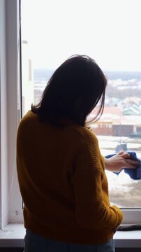 Brunette Woman Washes Window Pane Inside Apartment Using Blue Duster Cloth. Diligent Homemaker Scrubs Transparent Glass With Steady Movements, Keeping Interior Room Clean On Winter Morning.