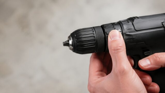 Close-up of a hand adjusting the chuck of a cordless drill against a blurred gray background, showcasing the tool's design and functionality in a workshop setting