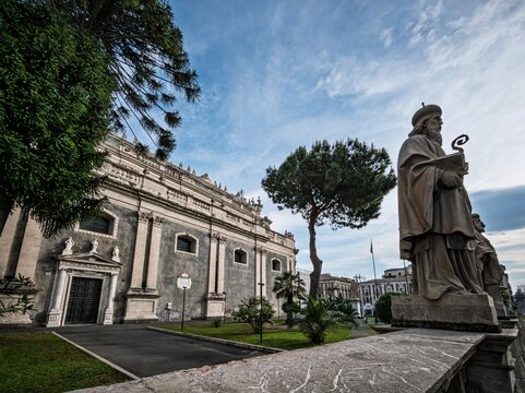 Side view of the majestic Catania Cathedral in Sicily, featuring grey lava stone walls, white limestone trim, and a historic saint statue in the foreground.