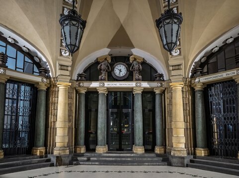 Symmetrical interior composition showcasing vaulted ceiling arches, dark marble columns, carved atlas clock, and hanging lanterns of Palazzo delle Poste in Catania.