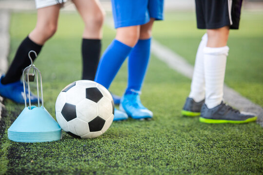 Selective focus to artificial green turf field and legs of young boys training football with a classic soccer ball and training cones equipment. Youth football practice day.