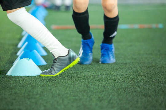 Selective focus to legs of young soccer players practicing footwork agility drills moving past blue training cones on an artificial green pitch. Youth football athletic drill and skills practice.
